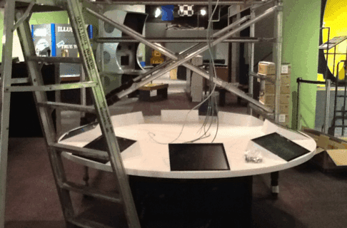 A circular table with eight digital touchscreens embedded around the surface. Ladders and scaffolding are on either side of the table as wires hang down from above.