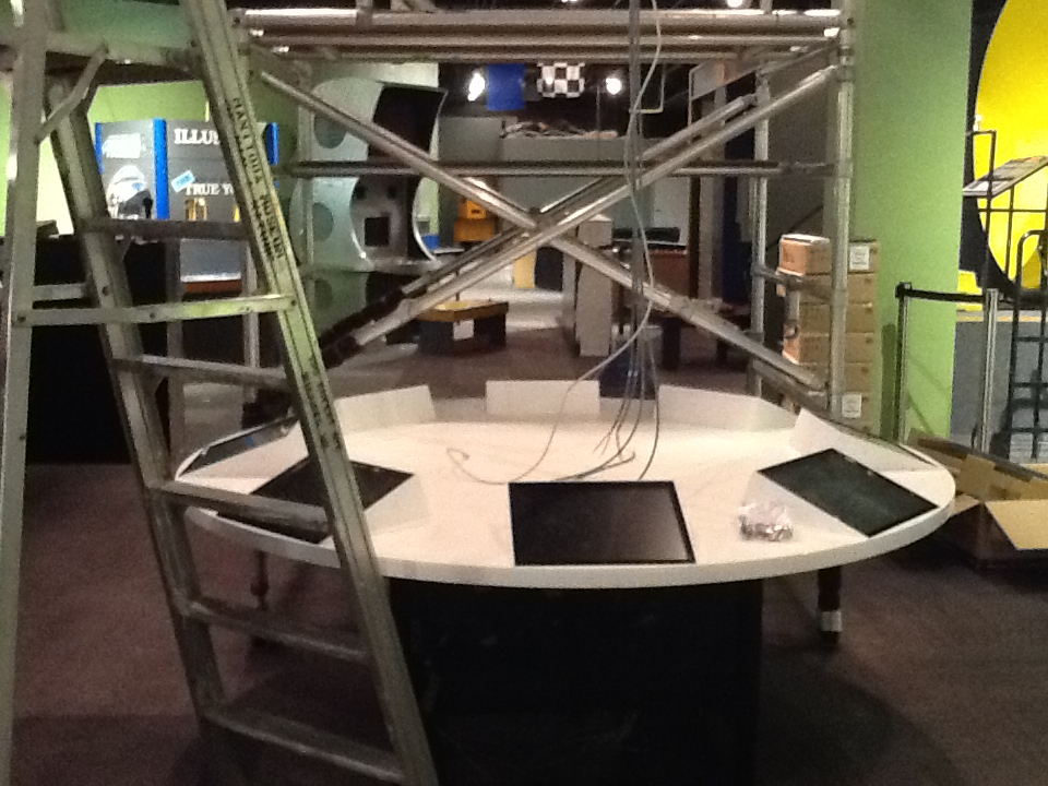 A circular table with eight digital touchscreens embedded around the surface. Ladders and scaffolding are on either side of the table as wires hang down from above.