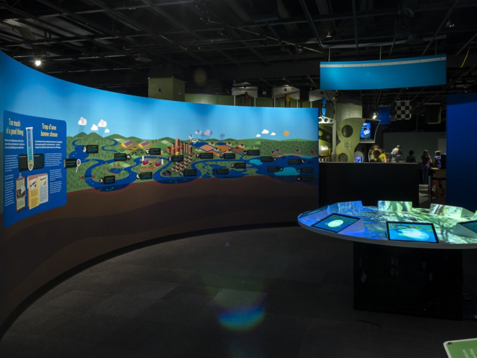 The Lake Winnipeg Shared Solutions exhibit in the Manitoba Museum Science Gallery. On the left, a curving divider wall displays a mural through the journey of water. On the right, a round table glows from eight embedded tablet screens.