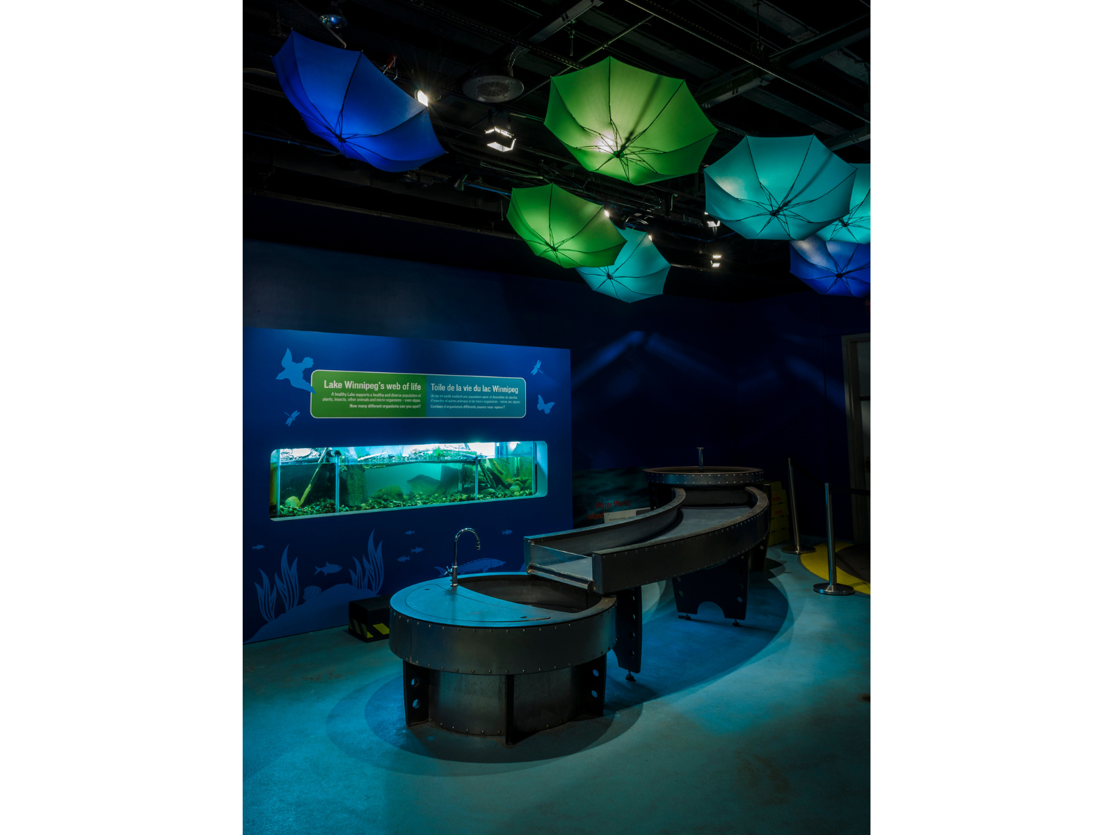 A sloping water table in front of a wall-embedded fish tank. Above the area blue and green umbrellas hang upside-down, lit by spotlights in the ceiling.
