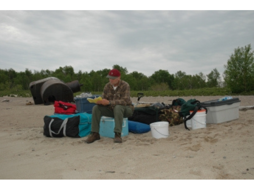 A man sitting with piles of luggage and packed boxes and bucks on a sandy beach.