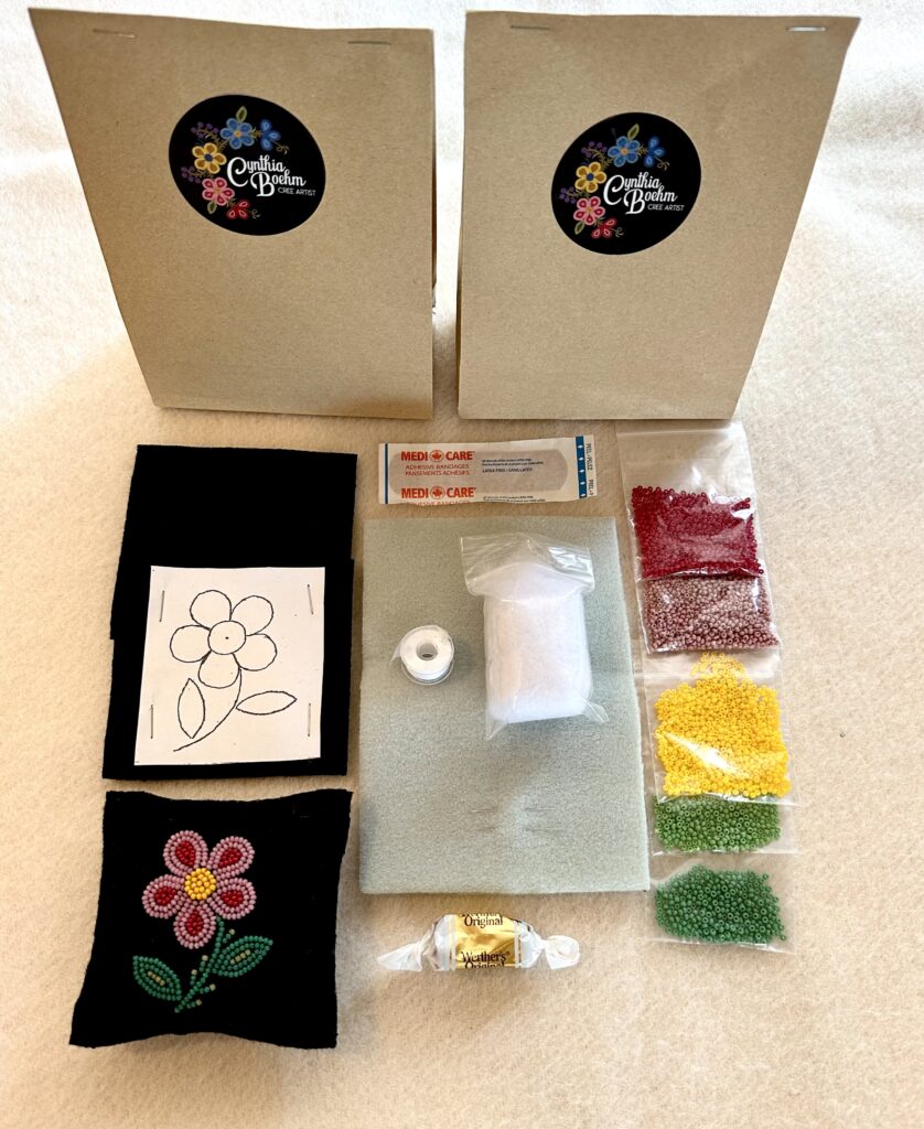 Two small paper bags (Beading Kits) sitting in front of the included supplies. Three bags of beads in red, yellow, and green, thread, needles, backing felt, a pattern of a flower, a band-aid, and a wrapped candy.