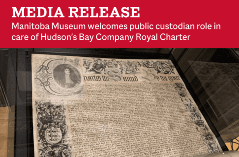 The HBC Charter on display in a glass case. A wide piece of parchment, still rolled at the bottom with intricate designs in the margins and the wording of the charter written with flourishes through the body of the page. On a red bar across the top text reads, "Media Release / Manitoba Museum welcomes public custodian role in care of Hudson's Bay Company Royal Charter".