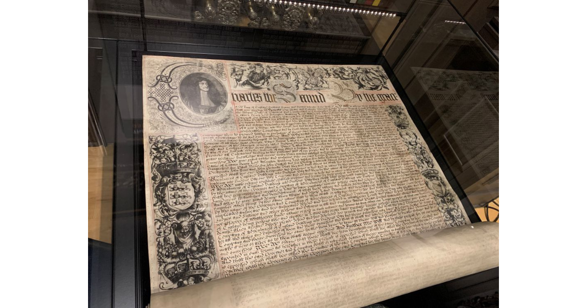 The HBC Charter on display in a glass case. A wide piece of parchment, still rolled at the bottom with intricate designs in the margins and the wording of the charter written with flourishes through the body of the page.