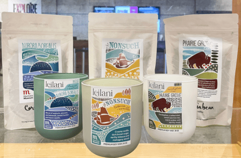 Three bags of loose lead tea from Cornelia Bean in Manitoba Museum themed flavours - Aurora Borealis, Nonsuch, and Prairie Grove. In front of the teas are three candles from Leilani Candle Collection in scents of the same themes.