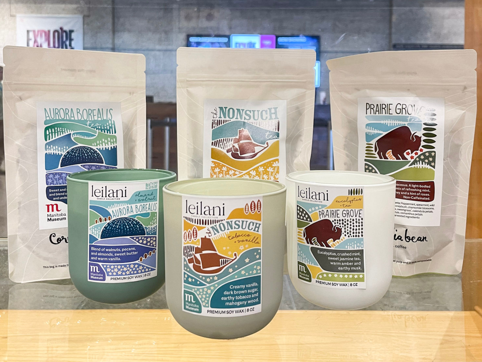 Three bags of loose lead tea from Cornelia Bean in Manitoba Museum themed flavours - Aurora Borealis, Nonsuch, and Prairie Grove. In front of the teas are three candles from Leilani Candle Collection in scents of the same themes.