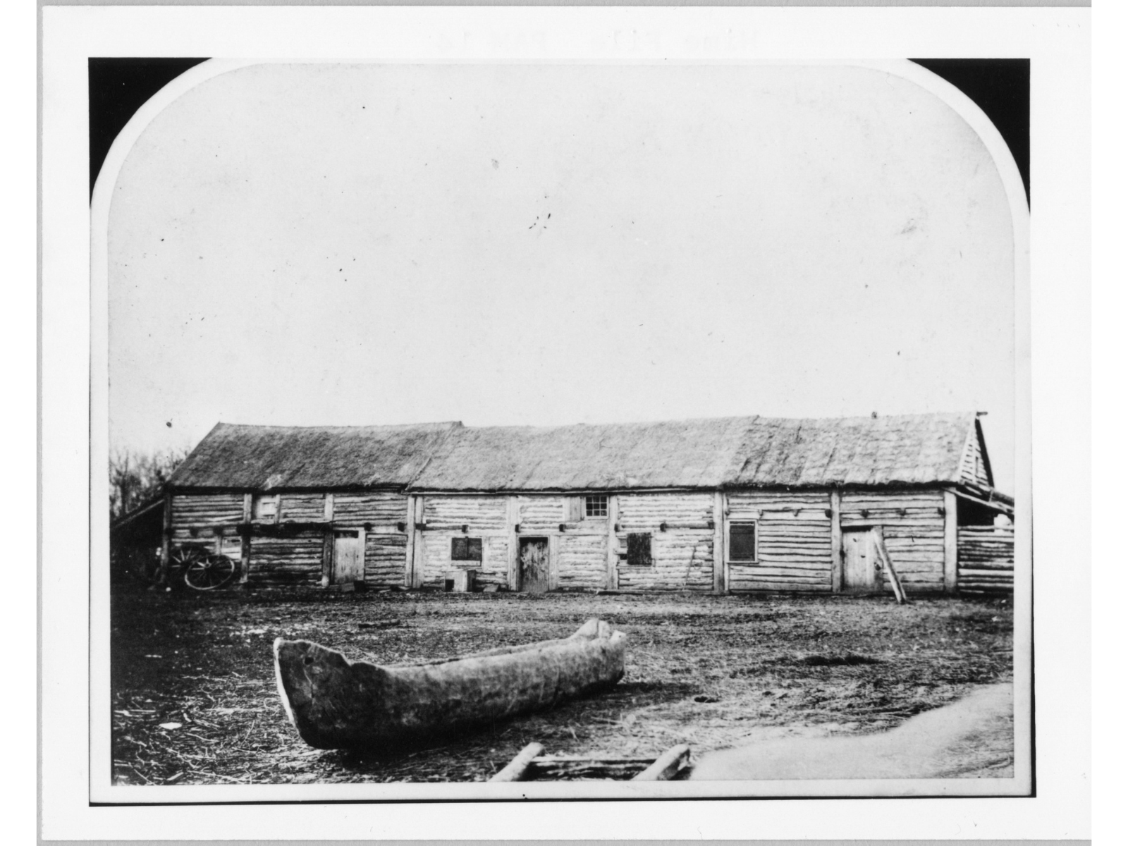Block and white photograph of a long, two-storied wooden building. In the foreground is a wooden canoe.