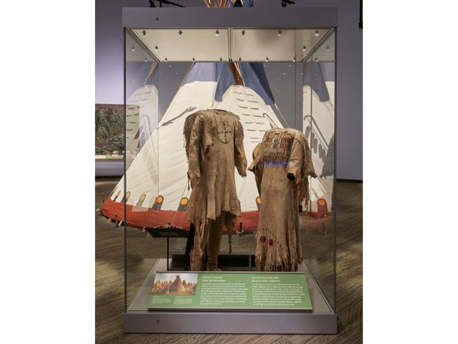 A display case with men's and women's Northern Plains style clothing on display. Through the glass part of a tipi can be seen.