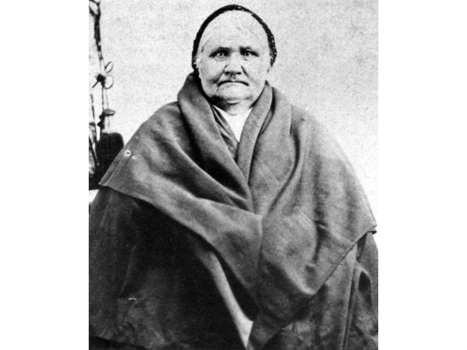 Black and white photograph of an older lady wearing a bulky shawl wrapped over her upper body and a kerchief over her white hair.