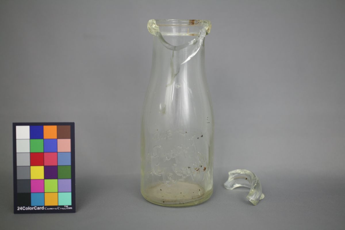 A wide necked glass bottle with a chunk broken off the top and laying beside the bottle.