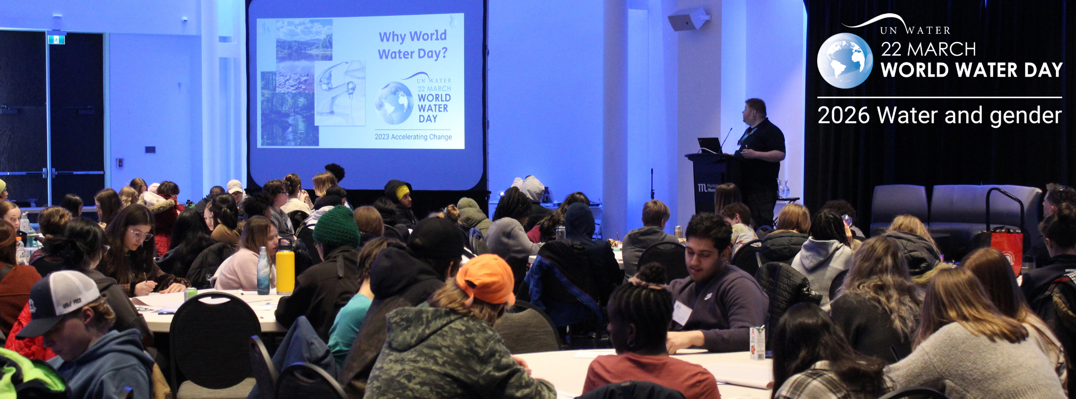 A large room with groups of students sitting around tables. In the back of the photo, a Museum staff person stands in front of a podium at the front of the room next to a presentation screen with 