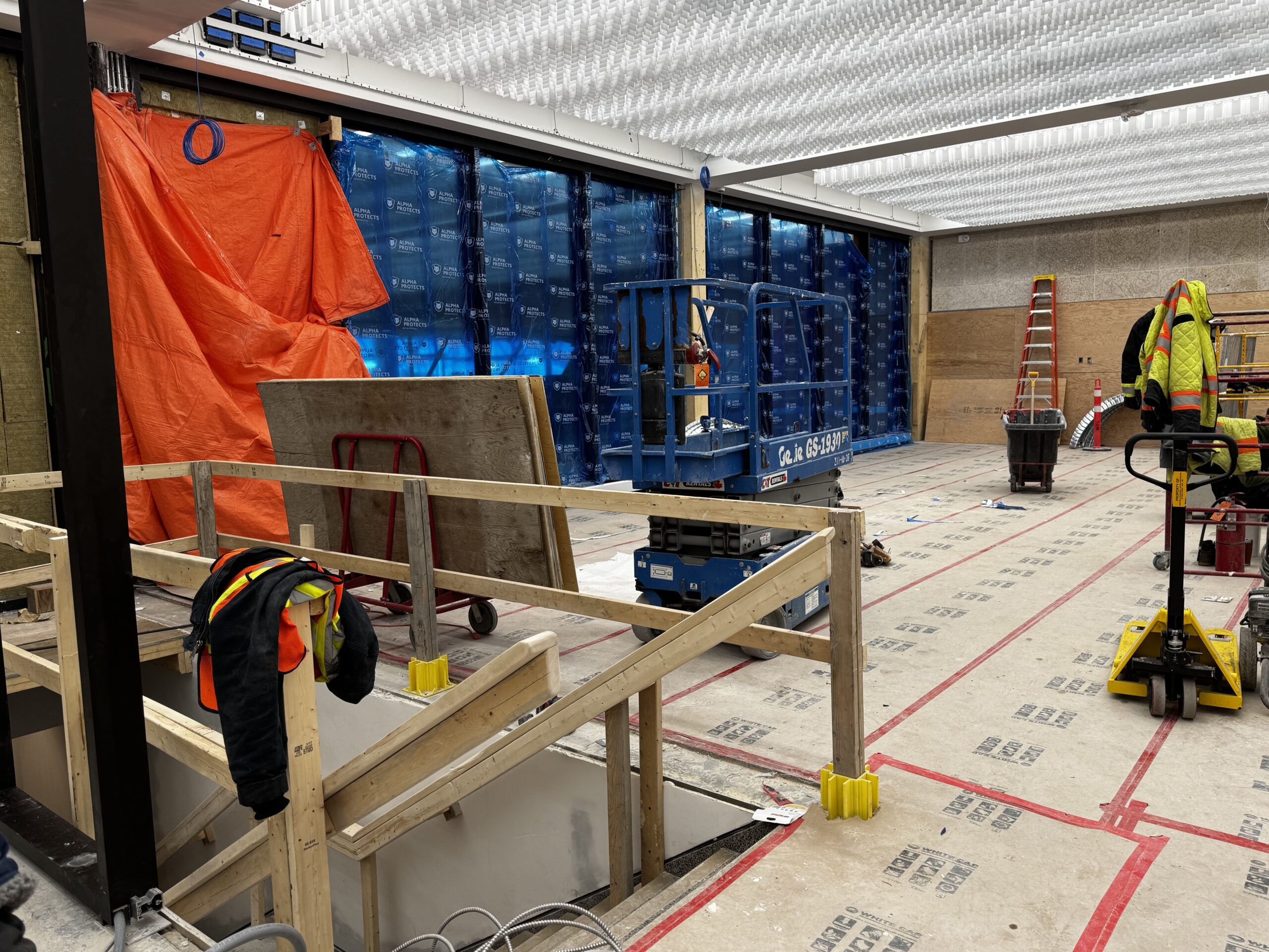 Construction site set up in the north half of the Manitoba Museum foyer. The exterior wall is of large glass windows with a blue protective covering on them, and temporary wooden railings have been set up in the stairwell going down to the Concourse.
