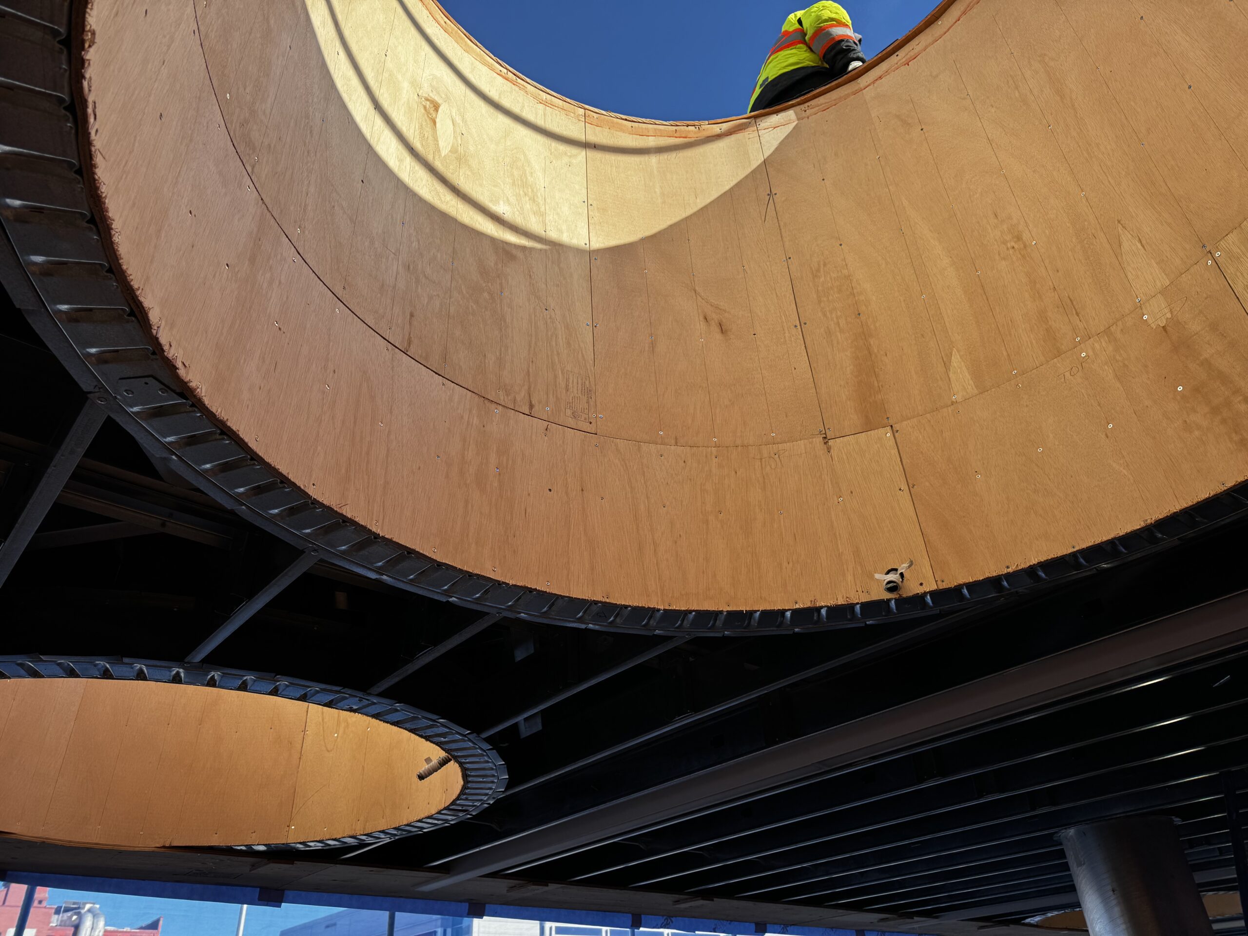 Looking up through a circular wooden framed skylight to a blue sky. An individual walks past above wearing a yellow high-vis jacket.