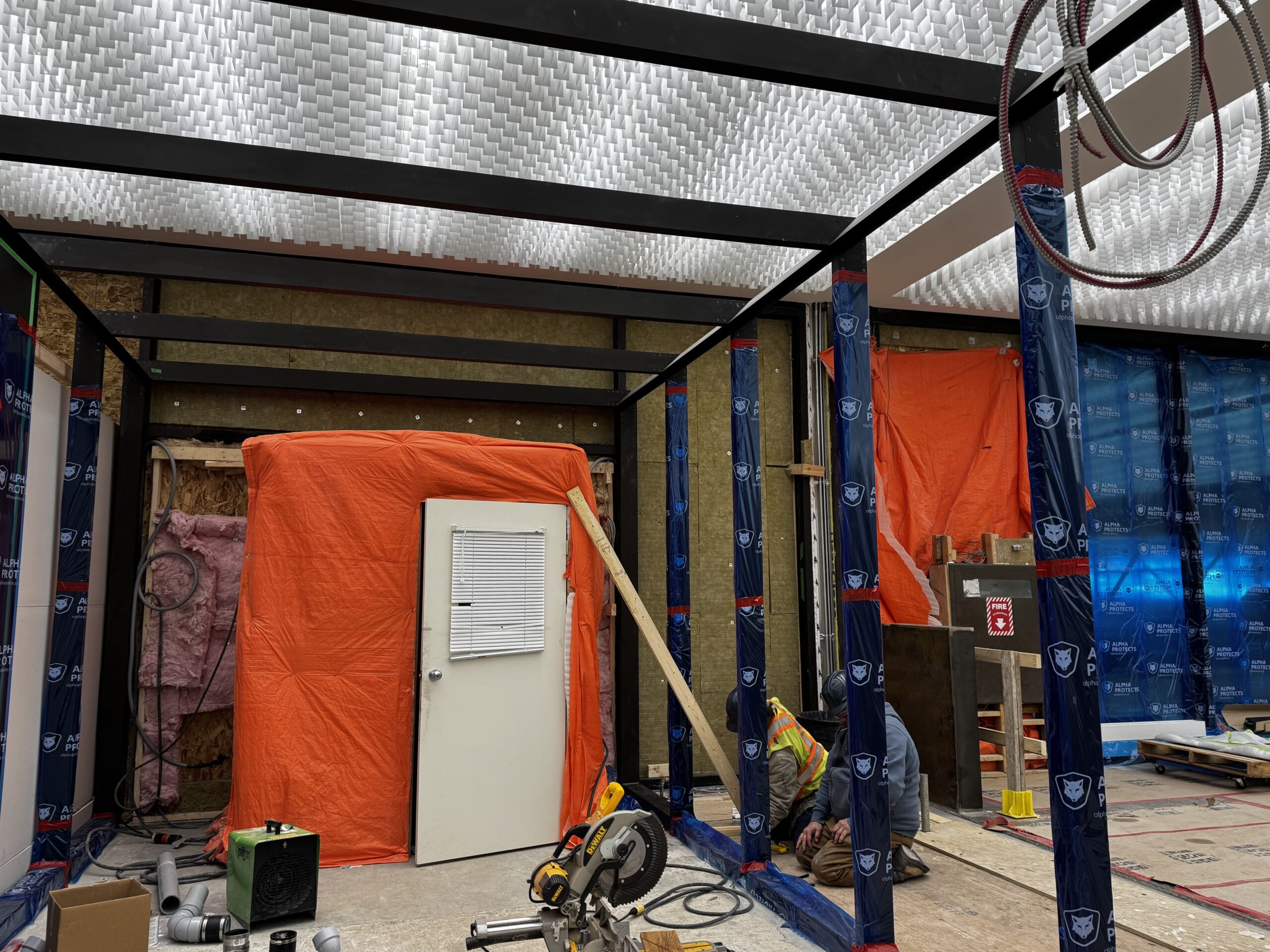 Metal framing over what will be the main entry-way. At the end is a orange-tarped area with a single door leading to the construction site.