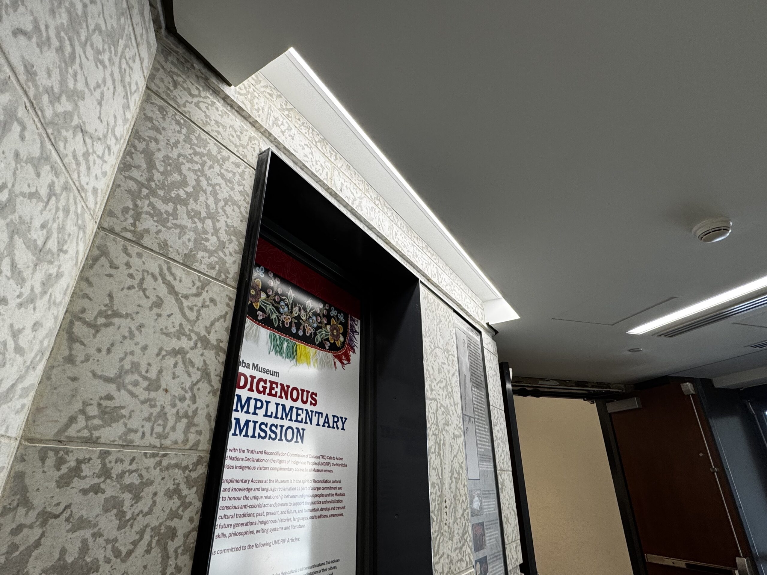 Looking up at a highlight light embedded in the ceiling, pointing towards a inset display frame with information about the Manitoba Museum's Indigenous Complimentary Admission.