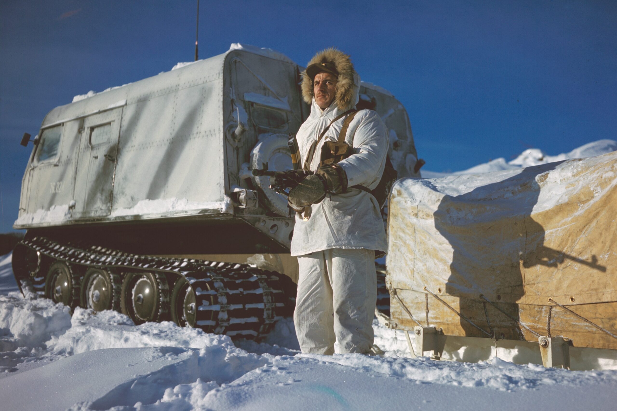 An individual standing behind a snowbank wearing a white fur-lined snowsuit holding a gun in front of a snow vehicle with large treads.