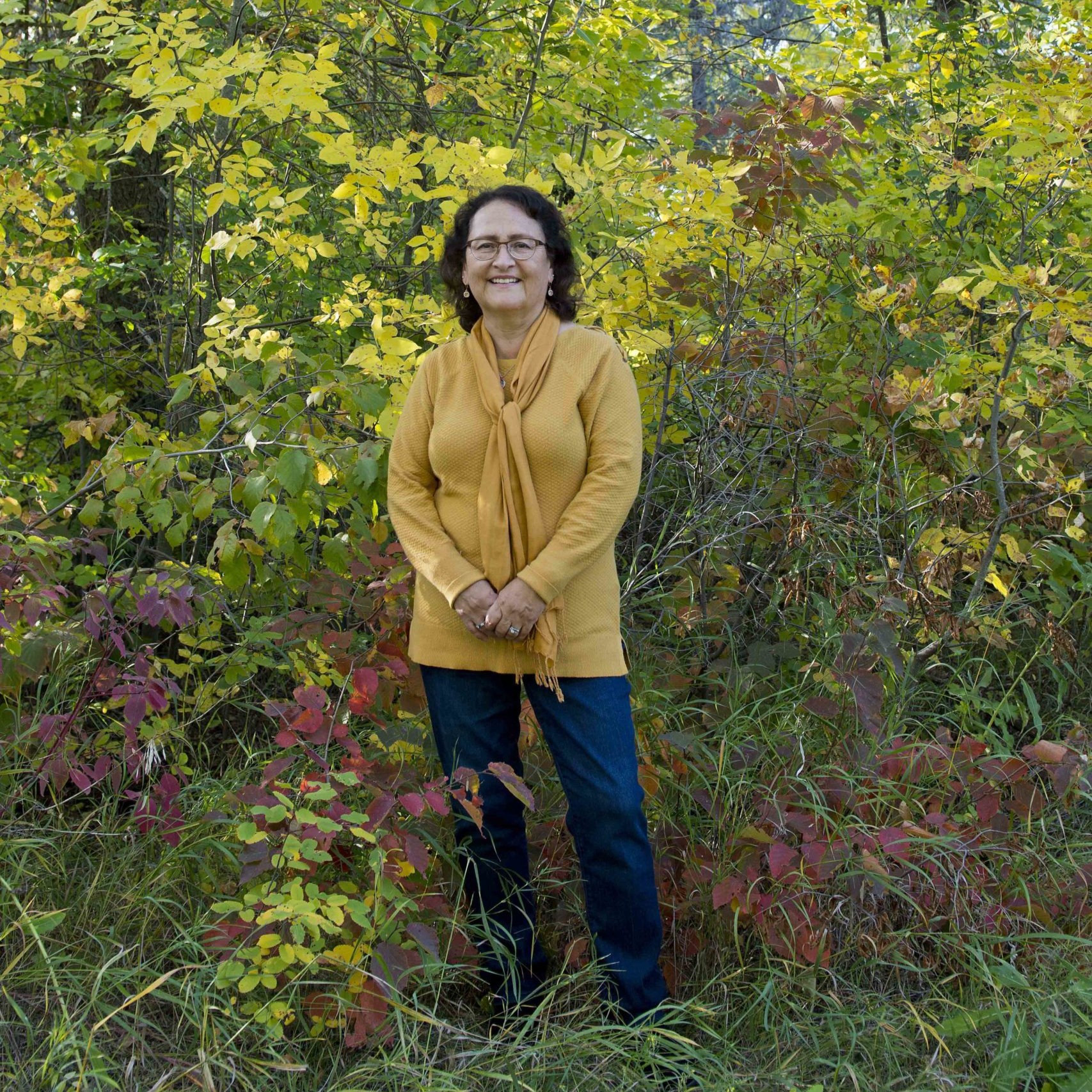 Elder Marlene Gallagher wearing a yellow top and blue jeans smiling for a photo while standing outside surrounded by autumn foliage.