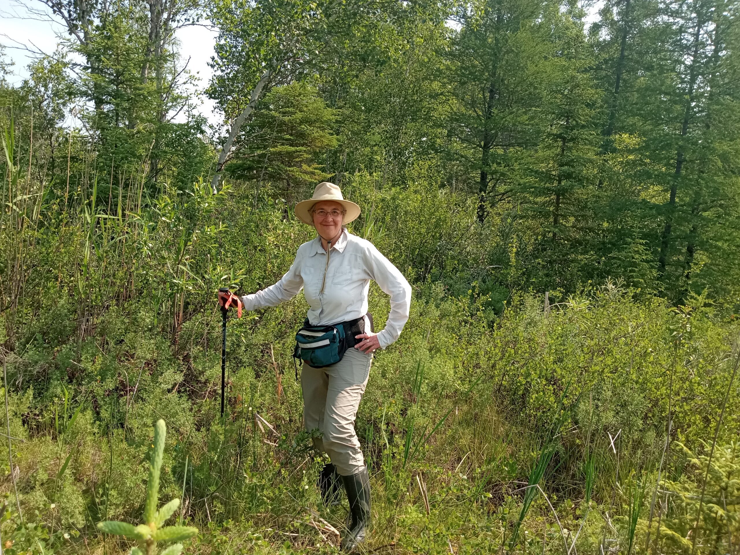 Woman in tall rubber boots with a wide-brimmed hat, large waist bag, and walking stick smiles for a photo among waist high brush with trees behind.