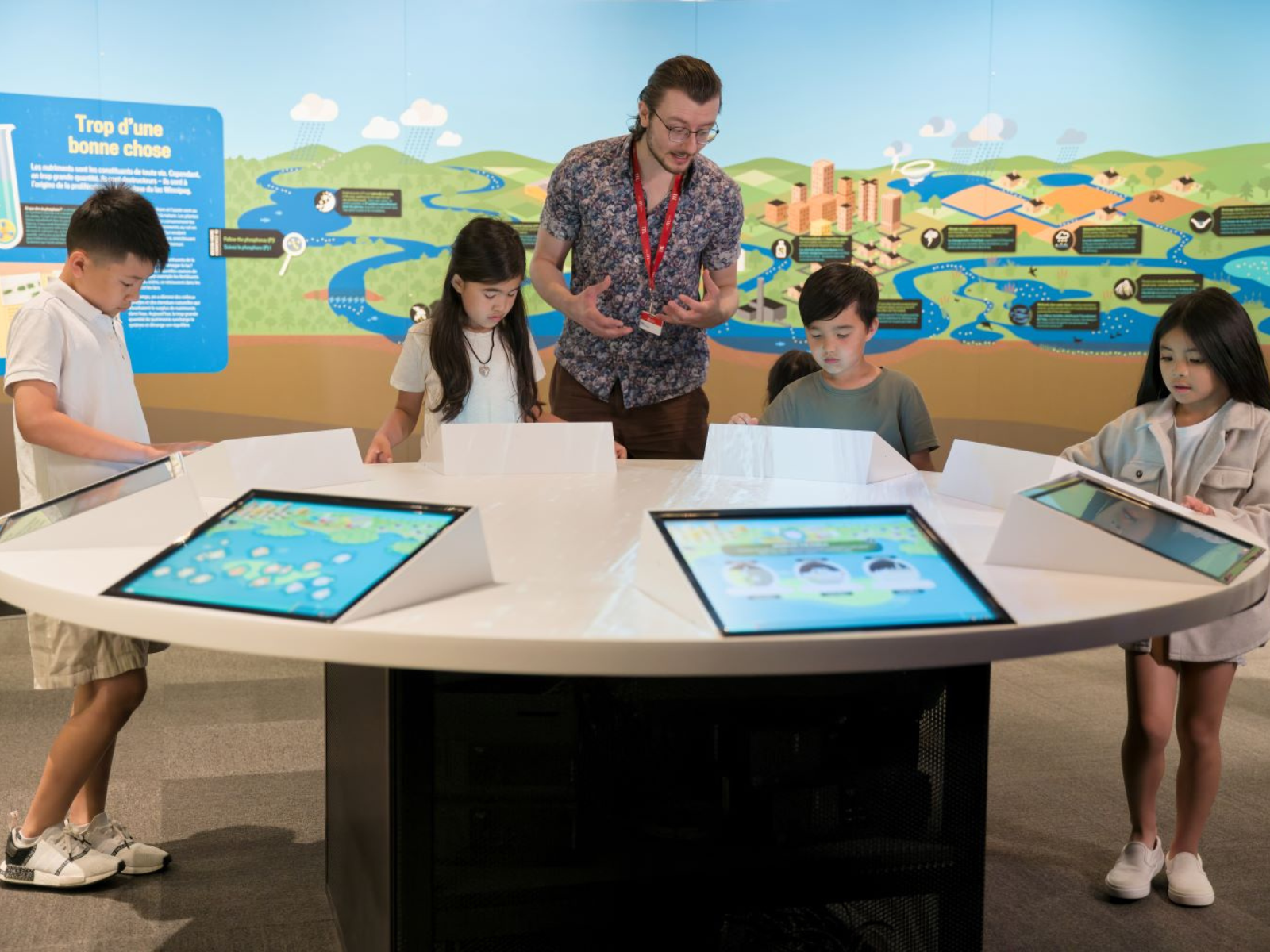 Four children standing around a round table engaging with exhibit material through with embedded digital screens. A museum staff member guides them through the activity.
