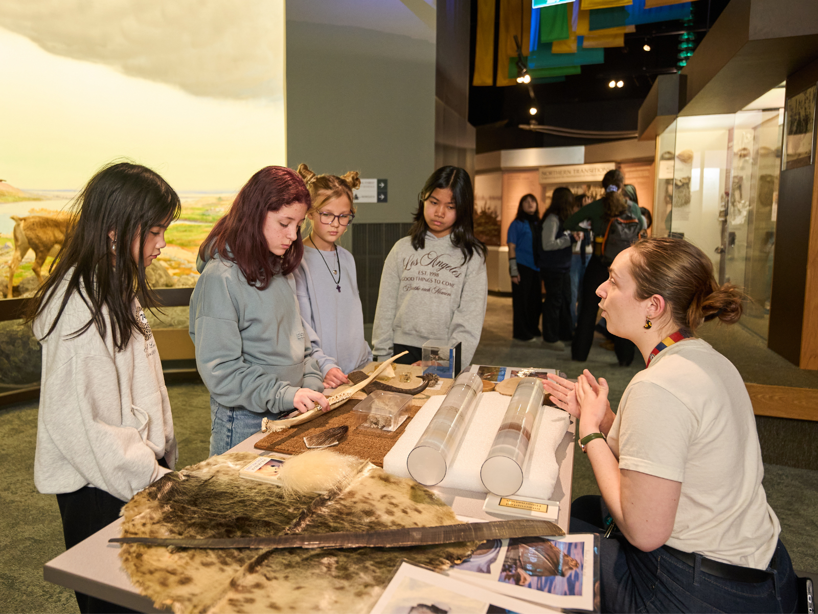 Four yoth stand in front of a pop-up display cart covered in fossils and specimens. A Museum staff member on the other side of the cart talks with them about the artifacts.