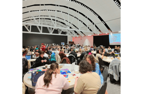 Looking out into a room with a high arched ceiling. Filling the room are people seated around round tables socializing and working on a variety of craft projects.