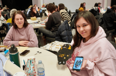 A smiling beader holds up a piece of beadwork and a mobile device with a inspiration picture on it. Another person seated at the same table smiles towards their beadwork.