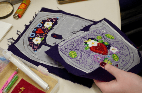 Close up on two pieces of beadwork in progress. One rests on the table and the other is held in hand as an out-of-frame beader works on it.