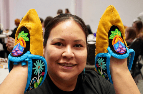 A smiling beader poses for a photo holding up a pair of moccasins on their hands.