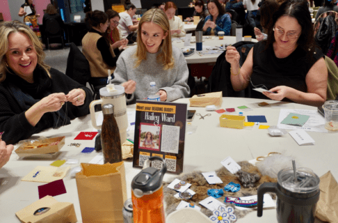 A group of three smiling people working on beadwork projects as they sit together at a table.
