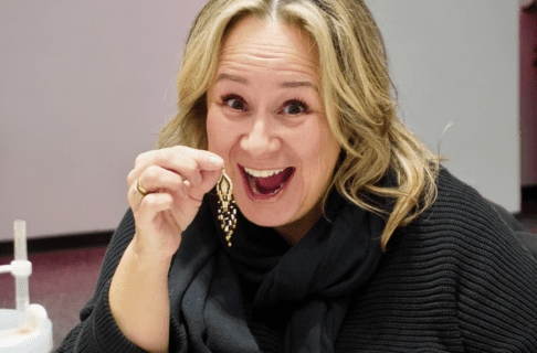 A beader smiles broadly as they hold up a detailed beaded earring.