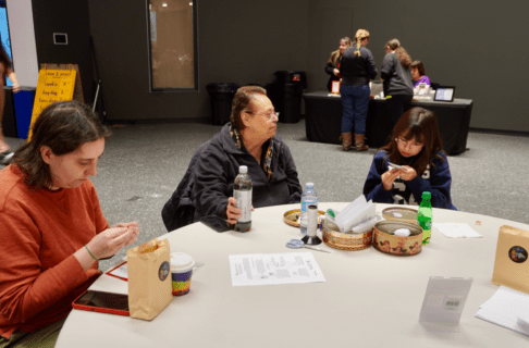Three people varying in age from child to older adult sitting together at a round table working on beadwork projects.