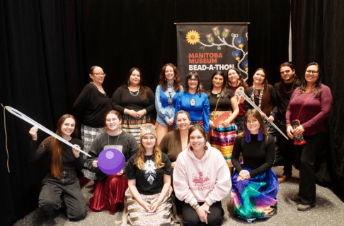 A group of fifteen people posing together for a group photo in front of a pop-up banner for the Manitoba Museum Bead-A-Thon. Several people are holding enlarged 2D props in the shape of needles and beads.