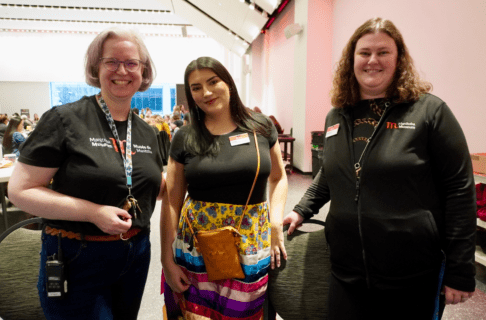 Three women smiling together for a photo. The woman on the left wears a Museum branded t-shirt, the woman in centre wears a Museum name tag on a black shirt with a colourful ribbon shirt, and the third woman on the right wears a Museum branded jacket and name tag.