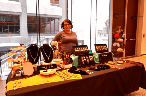 A person smiles to the camera from behind a table with displaying jewelry and beadwork.