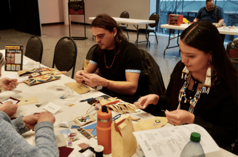 Two people sit side by side at a table each working on beadwork projects.