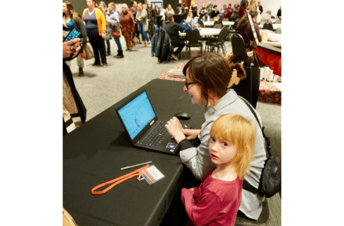 A woman sitting at a table with an open laptop in front of her and a child standing beside her. Across from the table is a line up of people waiting to check-in.