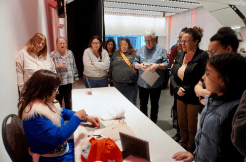 An individual sits at a table demonstrating a beading technique with a group of people standing around observing.