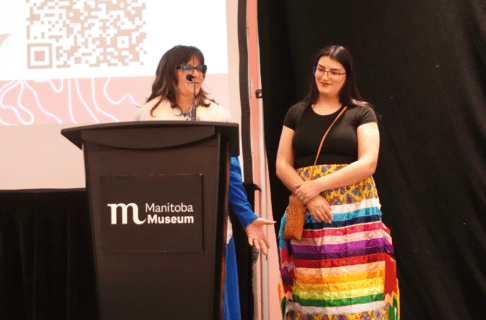 Two women standing together, one in front of a mic on a Manitoba Museum podium and the other, wearing a colourful ribbon skirt and black top, standing to the side.