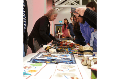 A Manitoba Museum staff person wearing gloves showing details of ancestral belongings on a table, with a group of people standings together looking at the belongings.