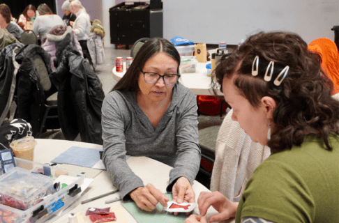 Two individuals sitting at a round table. One holds a piece of beadwork and leans forward pointing something out on it to the person seated next to them.