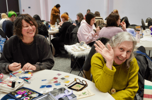 Two people seated at a round table in a room full of round places. The person on the right smiles and waves at the camera, as the person on the left smiles towards the first individual.