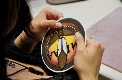 Looking over a beader's shoulder at the beadwork piece in their hand that they're working on.