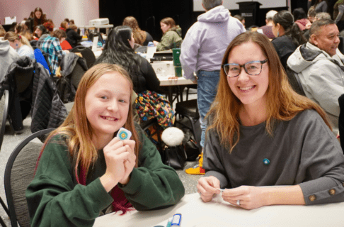 An adult and child smiling to the camera as they sit together at a table working on beadwork projects.