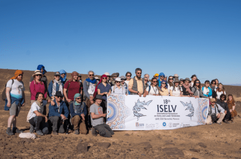 A group of more than three dozen people pose for a photo together in the desert with a large banner reading "18th ISELV International Symposium on Early and Lower Vertebrates / UHP, ESEF Berrechid, Morocco".