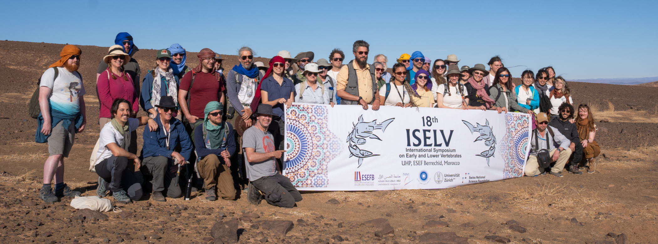 A group of more than three dozen people pose for a photo together in the desert with a large banner reading 
