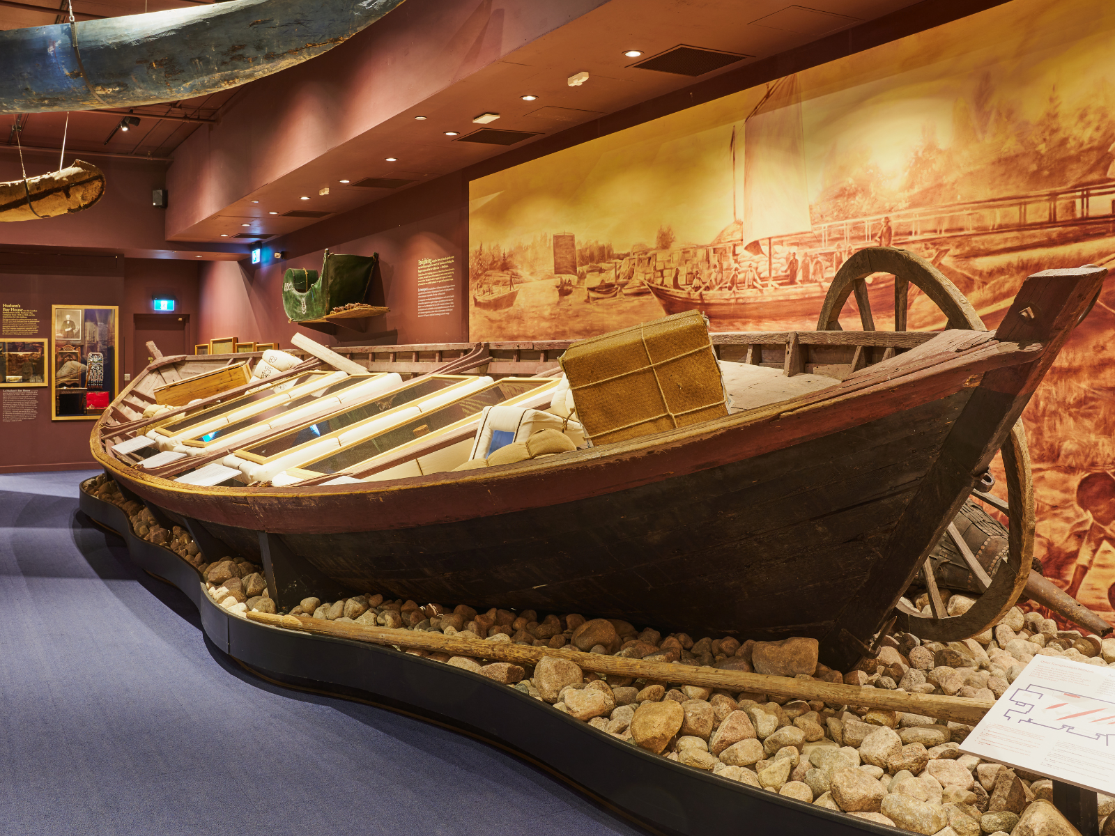 A large boat displayed in the Hudson's Bay Museum Collection Gallery with bales inside inlaid with screens containing digital content and artifact displays.