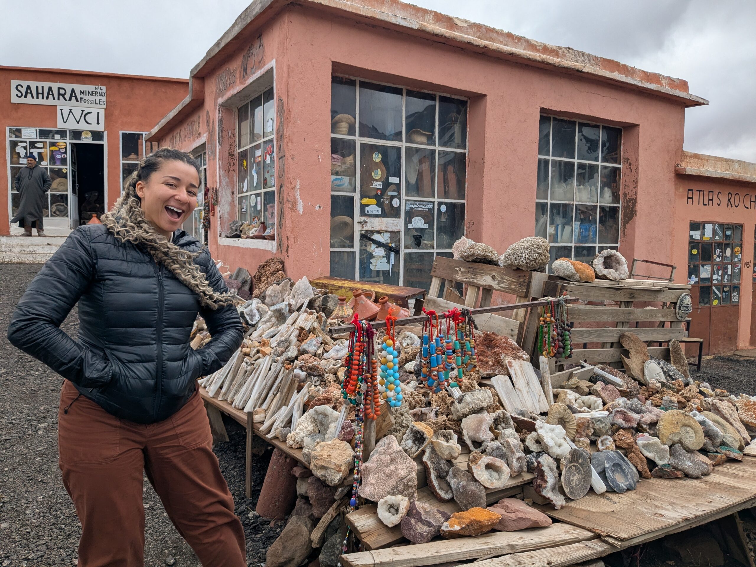 A smiling individual wearing a black puffer jacket poses smiling beside a table containing a variety of fossils and minerals outside a rosey coloured building.