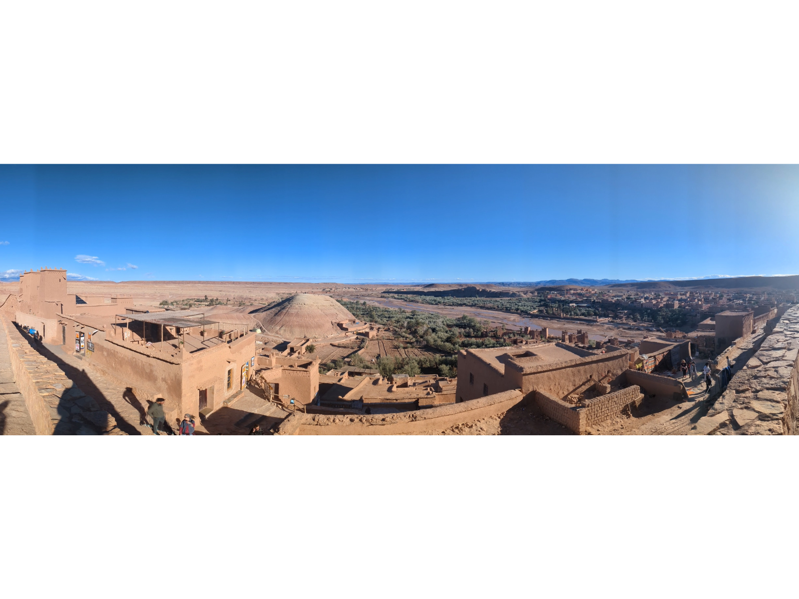 A panoramic view looking out from a high vantage over a fortified village built of red earthen clay. The desert as well as some treed areas along a riverbed stretch out in front of it.