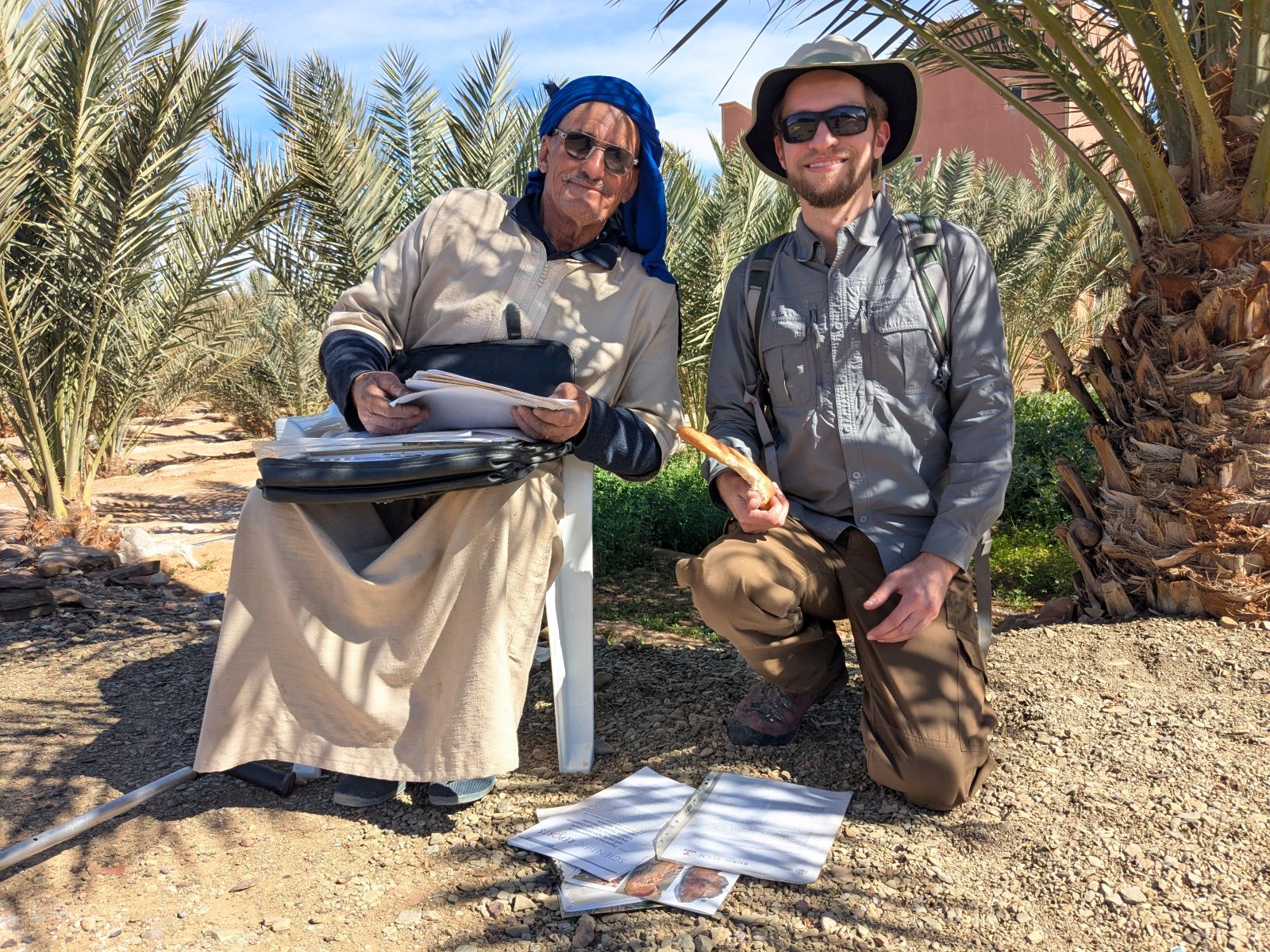 A photograph of two individuals posing for a photo together outside in a sandy environment near some leafy desert trees. Pictured left is Mohamed Oussaid Ben Moula and right is Dr. Joe Moysiuk.