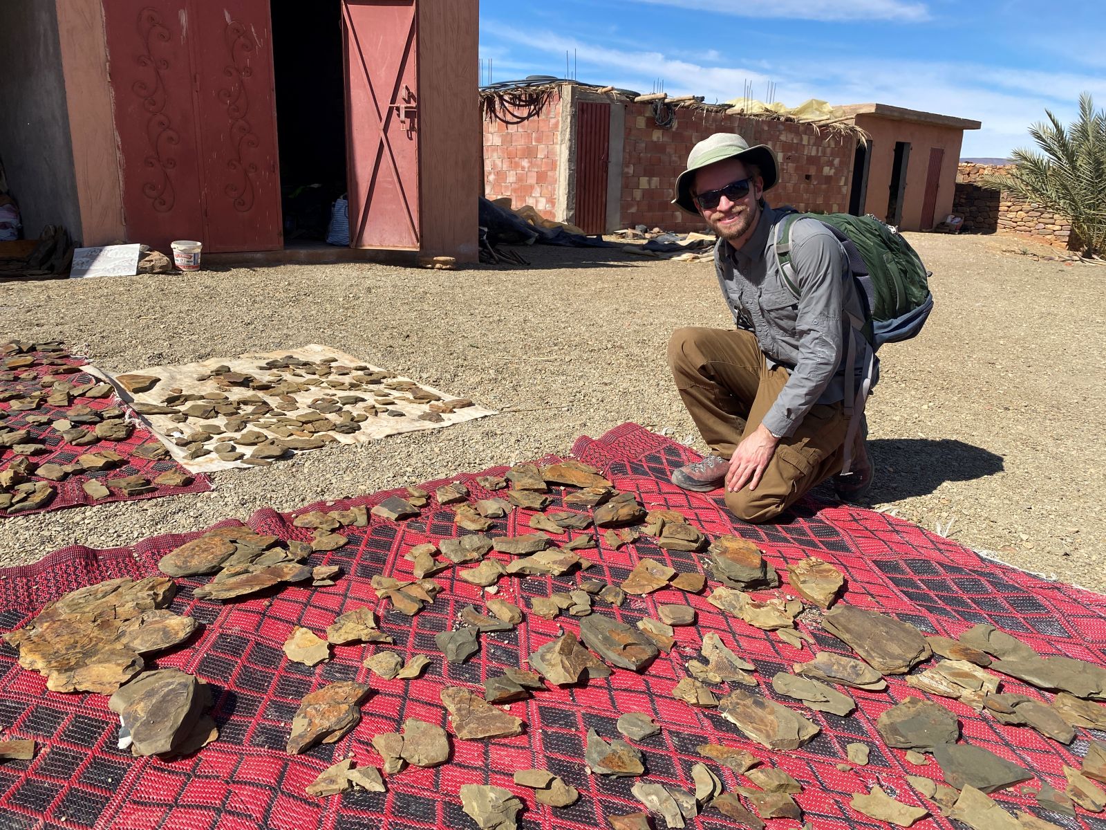 A dinividual posing for a photo kneeling beside a rug outside on a sandy ground. The rug is covered in a large selection of fossils.